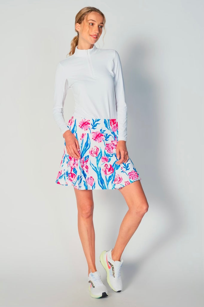 A woman stands confidently, facing camera, wears the G Lifestyle Flare Godet Skort in Tulip - floral print of the Spring Summer 2024 Collection. The skort flares out with playful, fluted edges, emphasizing movement and feminine silhouette. The skort appears to be a med-rise and the length hits her at mid-thigh. This skort is suitable for tennis, pickleball, padel tennis, golf.