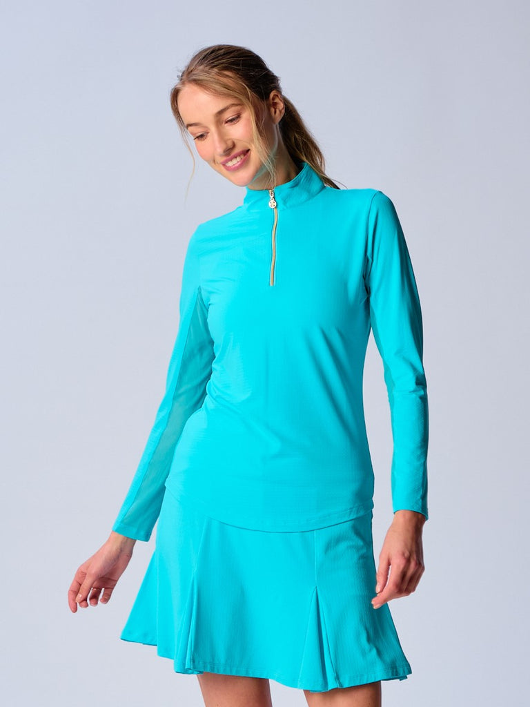 A woman wears the G Lifestyle Quarter Zip Sun Protection Top in Caribbean Turquoise. The top has long sleeves and an adjustable mock neck collar with a gold quarter zip, the brand’s logo discreetly placed on the right wrist. The fabric appears to have a lightweight, subtle texture, that likely offers menthol cooling, moisture wicking properties, breathability, sun safety and comfort. Suitable for various outdoor athletic activities such as golf, tennis, padel tennis, pickleball or even cycling.
