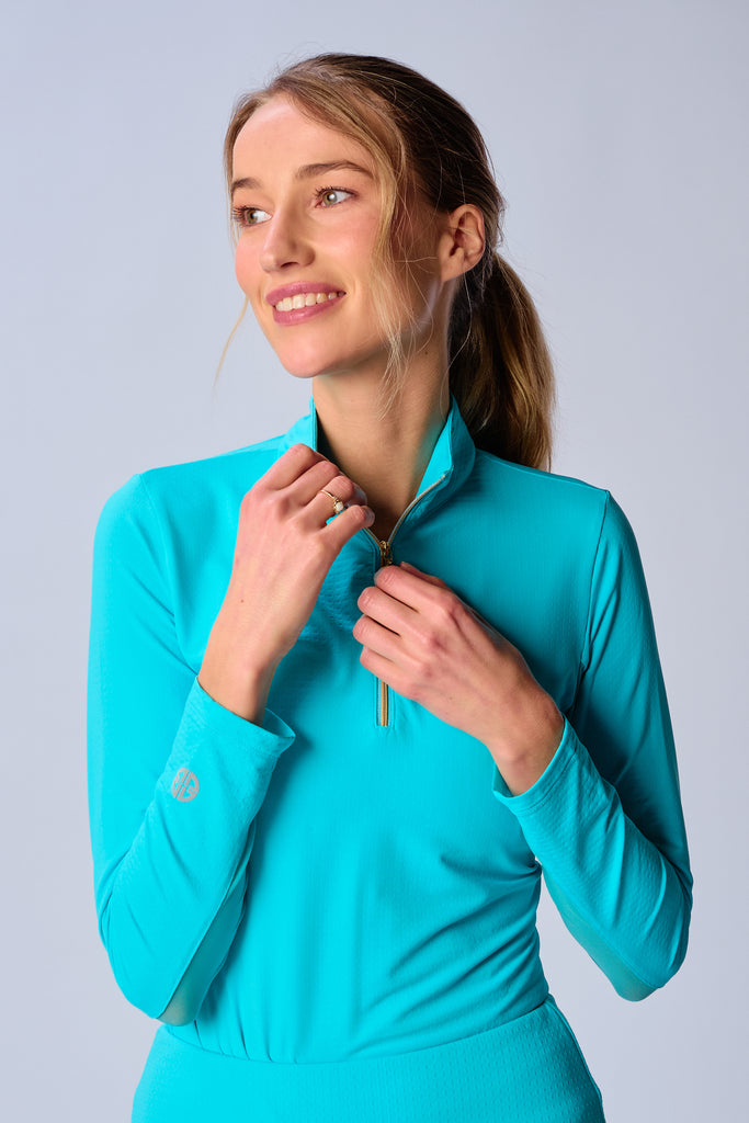 A close-up of a woman adjusting the collar of her G Lifestyle Quarter Zip Sun Protection Top in Caribbean Turquoise. The top is characterized by its high neckline and a prominent quarter-zip with a circular zipper pull bearing the brand’s logo, the brand’s logo discreetly placed on the right wrist as well. The zipper is gold-toned, adding a touch of luxury to the white top. The fabric has a texture, suggesting stretch and comfort, designed to offer sun protection. 