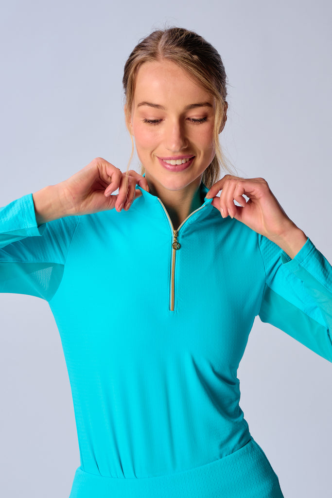 A close-up of a woman adjusting the collar of her G Lifestyle Quarter Zip Sun Protection Top in Caribbean Turquoise. The top is characterized by its high neckline and a prominent quarter-zip with a circular zipper pull bearing the brand’s logo. The zipper is gold-toned, adding a touch of luxury to the top. The fabric has a texture, suggesting stretch and comfort, designed to offer sun protection.