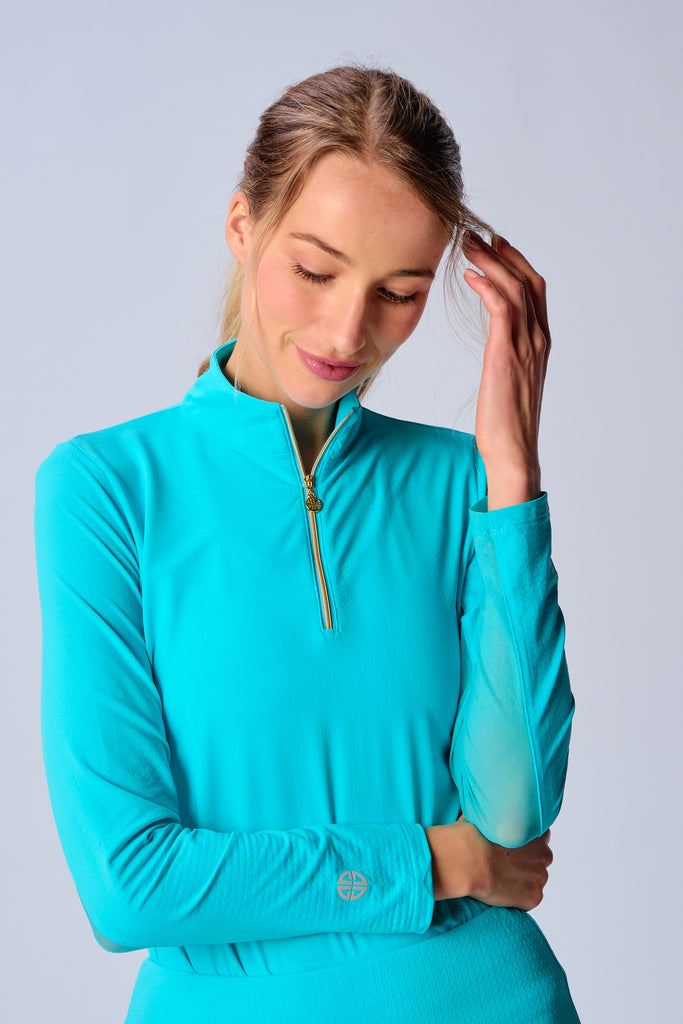 A close-up of a woman adjusting the collar of her G Lifestyle Quarter Zip Sun Protection Top in Caribbean Turquoise. The top is characterized by its high neckline and a prominent quarter-zip with a circular zipper pull bearing the brand’s logo, the brand’s logo discreetly placed on the right wrist as well. The zipper is gold-toned, adding a touch of luxury to the top. The fabric has a texture, suggesting stretch and comfort, designed to offer sun protection. 