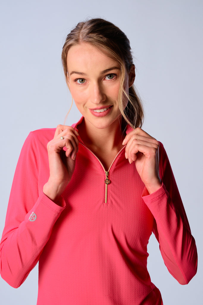 A close-up of a woman adjusting the collar of her G Lifestyle Quarter Zip Sun Protection Top in Coral. The top is characterized by its high neckline and a prominent quarter-zip with a circular zipper pull bearing the brand’s logo, the brand’s logo discreetly placed on the right wrist as well. The zipper is gold-toned, adding a touch of luxury to the white top. The fabric has a texture, suggesting stretch and comfort, designed to offer sun protection. 