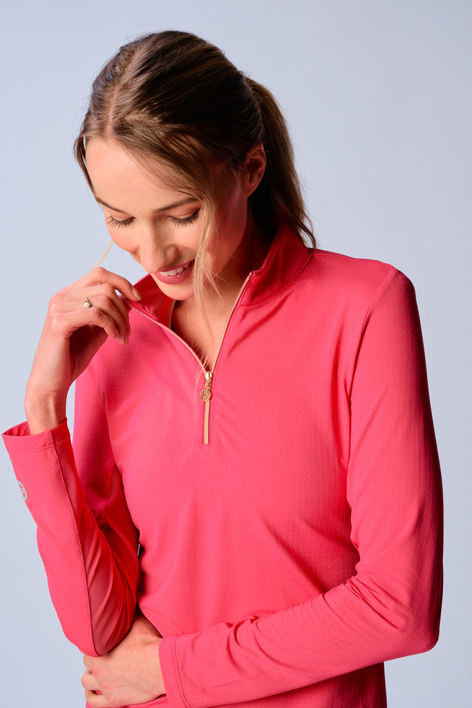 A close-up of a woman adjusting the collar of her G Lifestyle Quarter Zip Sun Protection Top in Coral. The top is characterized by its high neckline and a prominent quarter-zip with a circular zipper pull bearing the brand’s logo, the brand’s logo discreetly placed on the right wrist as well. The zipper is gold-toned, adding a touch of luxury to the top. The fabric has a texture, suggesting stretch and comfort, designed to offer sun protection. 
