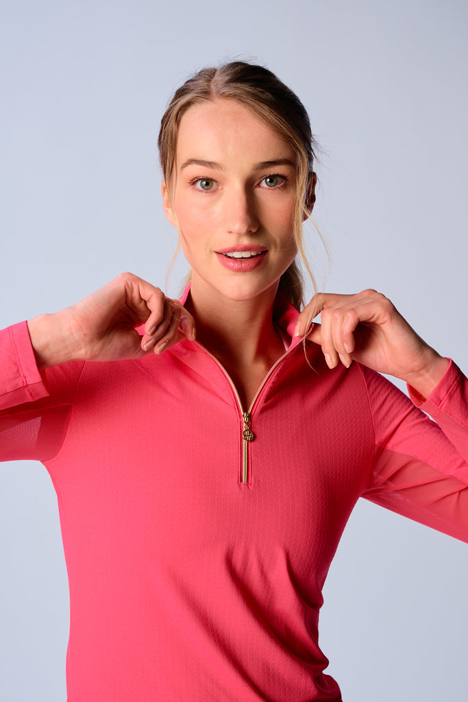 A close-up of a woman adjusting the collar of her G Lifestyle Quarter Zip Sun Protection Top in Coral. The top is characterized by its high neckline and a prominent quarter-zip with a circular zipper pull bearing the brand’s logo, the brand’s logo discreetly placed on the right wrist as well. The zipper is gold-toned, adding a touch of luxury to the top. The fabric has a texture, suggesting stretch and comfort, designed to offer sun protection. 
