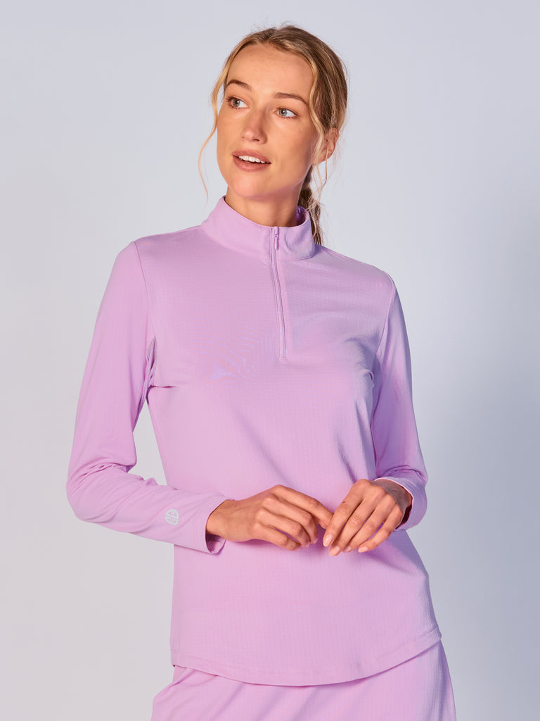 A woman wears G Lifestyle Quarter Zip Sun Protection Top in Lavender. The top has long sleeves, an adjustable mock neck collar with a flat quarter zip, the brand’s logo discreetly placed on the right wrist. The fabric appears to have a lightweight, subtle texture, that likely offers menthol cooling, moisture wicking, breathability, sun safety and comfort. Suitable for golf, tennis, padel tennis, pickleball or even cycling.