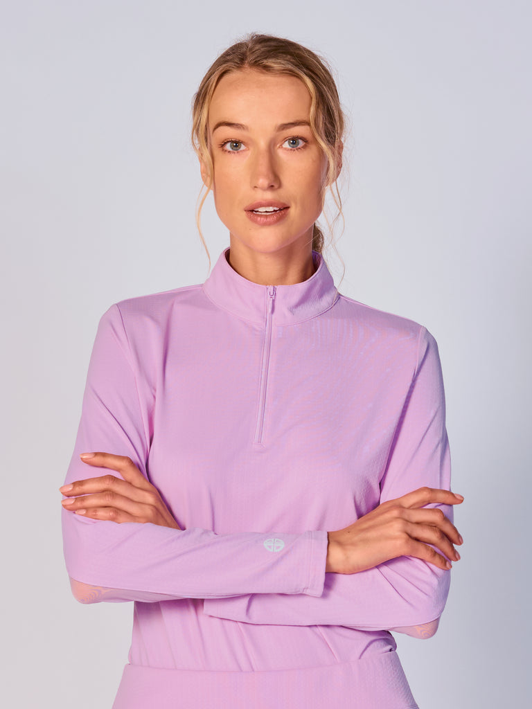 A woman wears G Lifestyle Quarter Zip Sun Protection Top in Lavender. The top has long sleeves, an adjustable mock neck collar with a flat quarter zip, the brand’s logo discreetly placed on the right wrist. The fabric appears to have a lightweight, subtle texture, that likely offers menthol cooling, moisture wicking, breathability, sun safety and comfort. Suitable for golf, tennis, padel tennis, pickleball or even cycling.
