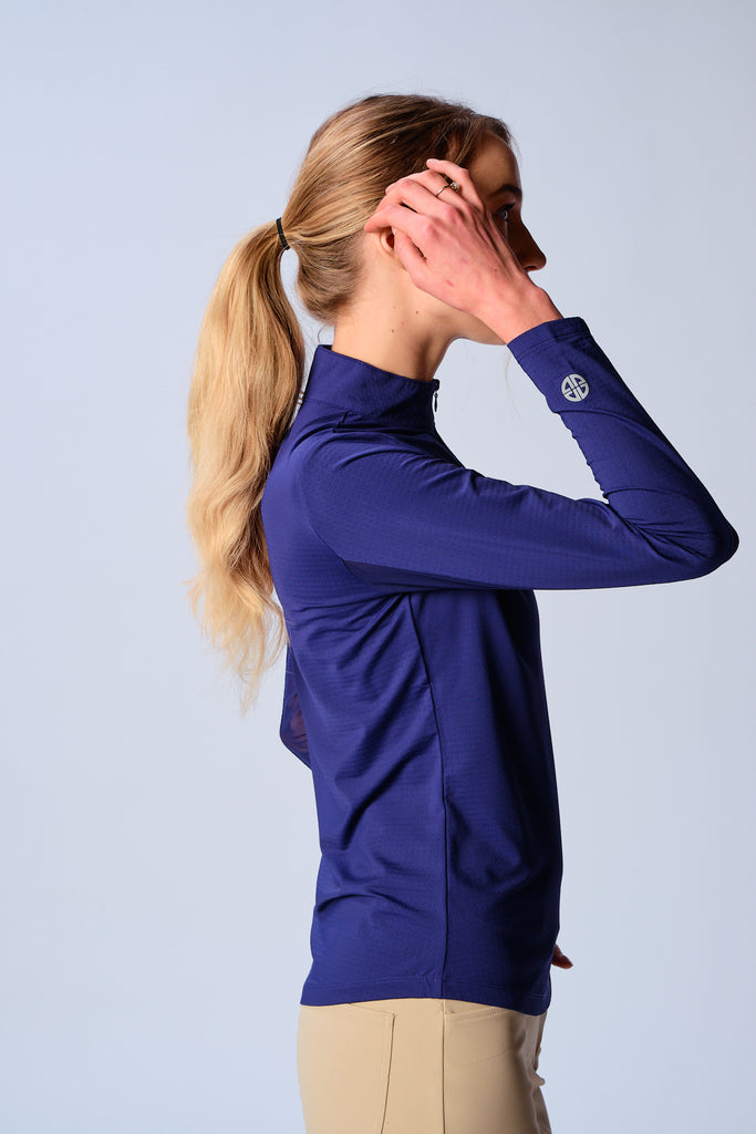 A woman, featured from the right side, wears the G Lifestyle Quarter Zip Sun Protection Top in True Navy. The top has long sleeves, an adjustable mock neck collar with a flat quarter zip, the brand’s logo discreetly placed on the right wrist. The fabric appears to have a lightweight, subtle texture, that likely offers menthol cooling, moisture wicking, breathability, sun safety and comfort. Suitable for golf, tennis, padel tennis, pickleball or even cycling.