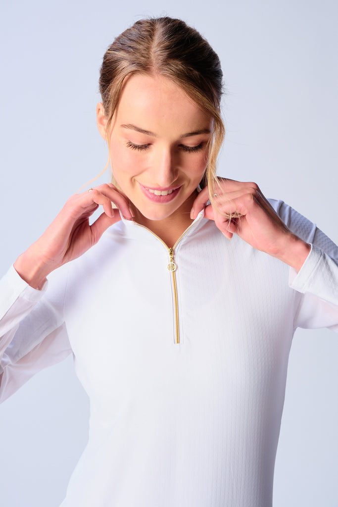 A close-up of a woman adjusting the collar of her G Lifestyle Quarter Zip Sun Protection Top in White. The top is characterized by its high neckline and a prominent quarter-zip with a circular zipper pull bearing the brand’s logo. The zipper is gold-toned, adding a touch of luxury to the white top. The fabric has a texture, suggesting stretch and comfort, designed to offer sun protection. 
