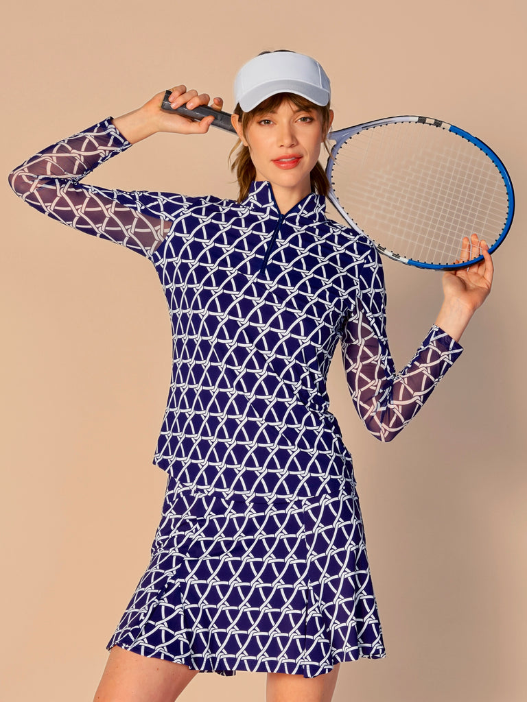 A woman is holding a tennis racket, showcasing a G Lifestyle Quarter Zip Sun Protection Top in Nautical True Navy – marine-inspired rope-knot print from Spring Summer 2024 Collection. The top has long sleeves, an adjustable mock neck collar with a flat quarter zip. The fabric appears to have a lightweight, subtle texture, that likely offers menthol cooling, moisture wicking, breathability, sun safety and comfort. Suitable for golf, tennis, padel tennis, pickleball or even cycling.