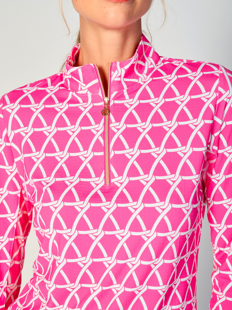 A close-up of a woman adjusting the collar of her G Lifestyle Quarter Zip Sun Protection Top in Nautical Hot Pink – marine-inspired rope-knot print from Spring Summer 2024 Collection. The top is characterized by its high neckline and a prominent quarter-zip with a circular zipper pull bearing the brand’s logo. The zipper is gold-toned, adding a touch of luxury to the top. The fabric has a texture, suggesting stretch and comfort, designed to offer sun protection