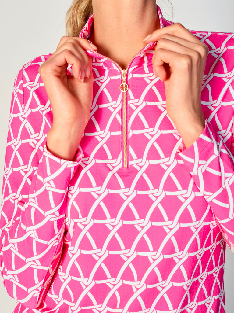 A close-up of a woman adjusting the collar of her G Lifestyle Quarter Zip Sun Protection Top in Nautical Hot Pink – marine-inspired rope-knot print from Spring Summer 2024 Collection. The top is characterized by its high neckline and a prominent quarter-zip with a circular zipper pull bearing the brand’s logo. The zipper is gold-toned, adding a touch of luxury to the top. The fabric has a texture, suggesting stretch and comfort, designed to offer sun protection