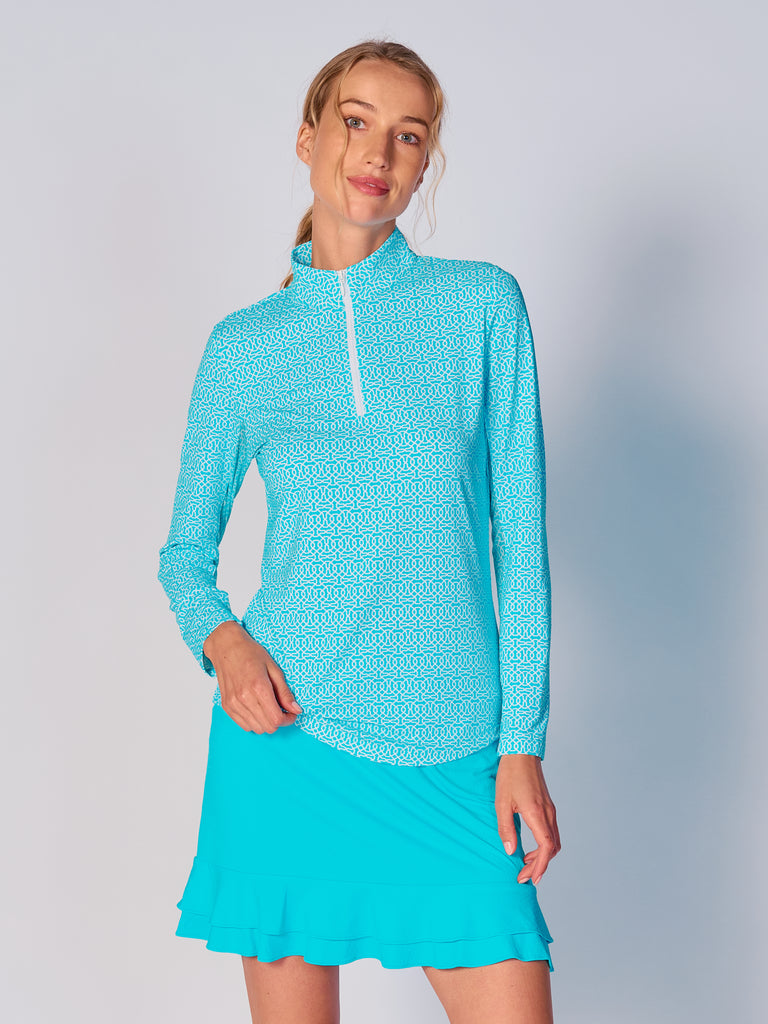 A woman wears the G Lifestyle Quarter Zip Sun Protection Top in Cubic Caribbean Turquoise - geometric print of the Spring Summer 2024 Collection. The top has long sleeves, an adjustable mock neck collar with a flat quarter zip. The fabric appears to have a lightweight, subtle texture, that likely offers menthol cooling, moisture wicking, breathability, sun safety and comfort. Suitable for golf, tennis, padel tennis, pickleball or even cycling.