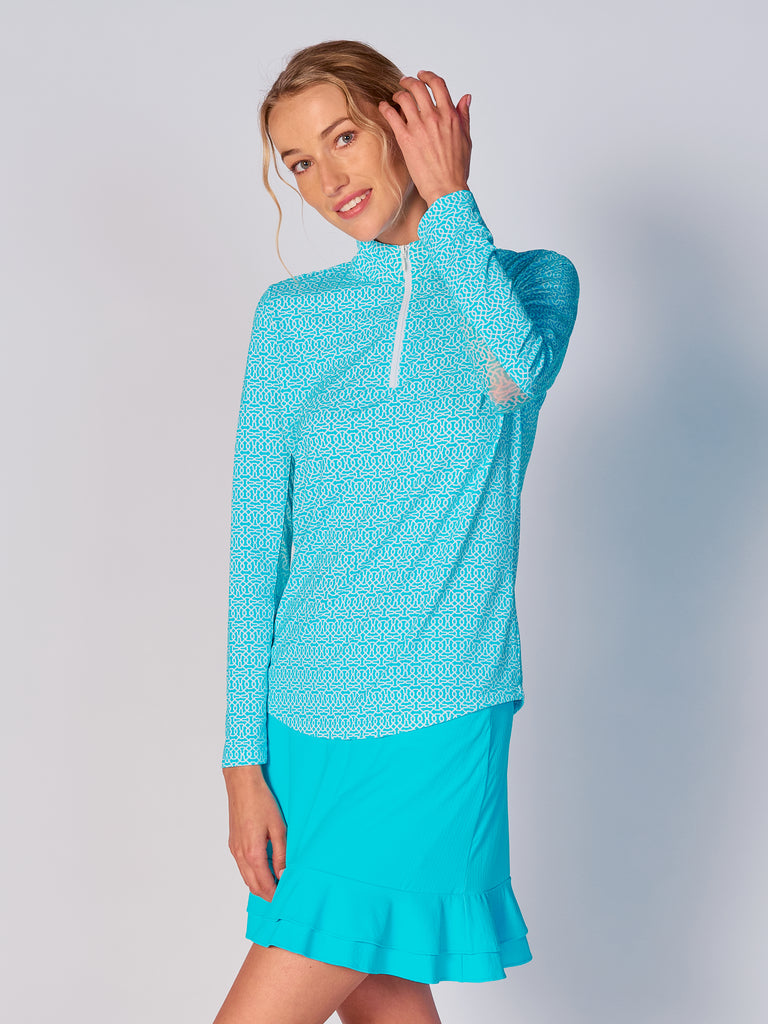 A woman wears the G Lifestyle Quarter Zip Sun Protection Top in Cubic Caribbean Turquoise - geometric print of the Spring Summer 2024 Collection. The top has long sleeves, an adjustable mock neck collar with a flat quarter zip. The fabric appears to have a lightweight, subtle texture, that likely offers menthol cooling, moisture wicking, breathability, sun safety and comfort. Suitable for golf, tennis, padel tennis, pickleball or even cycling.