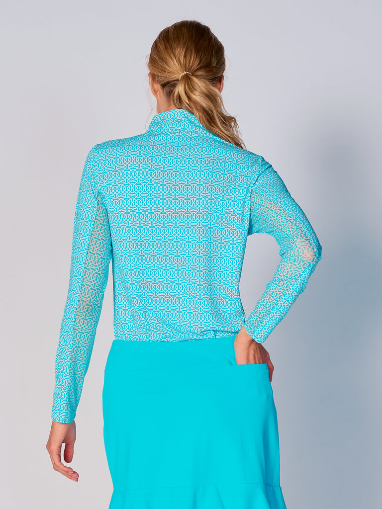 A woman is showcasing the back of the G Lifestyle Quarter Zip Sun Protection Top Cubic Caribbean Turquoise - geometric print of the Spring Summer 2024 Collection. The top is tailored with a form-fitting silhouette, complete with long sleeves and a high collar for maximum sun protection. Notably, it includes smooth mesh underarm inserts, which are seamlessly integrated for enhanced breathability, ensuring comfort during active use. 