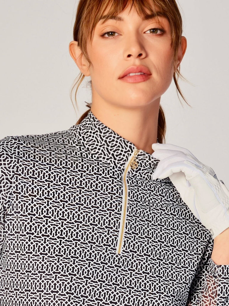 A close-up of a woman adjusting the collar of her G Lifestyle Quarter Zip Sun Protection Top in Cubic Black - brand’s geometric print of the Spring Summer 2024 Collection. The top is characterized by its high neckline and a prominent quarter-zip with a circular zipper pull bearing the brand’s logo. The zipper is gold-toned, adding a touch of luxury to the top. The fabric has a texture, suggesting stretch and comfort, designed to offer sun protection. 