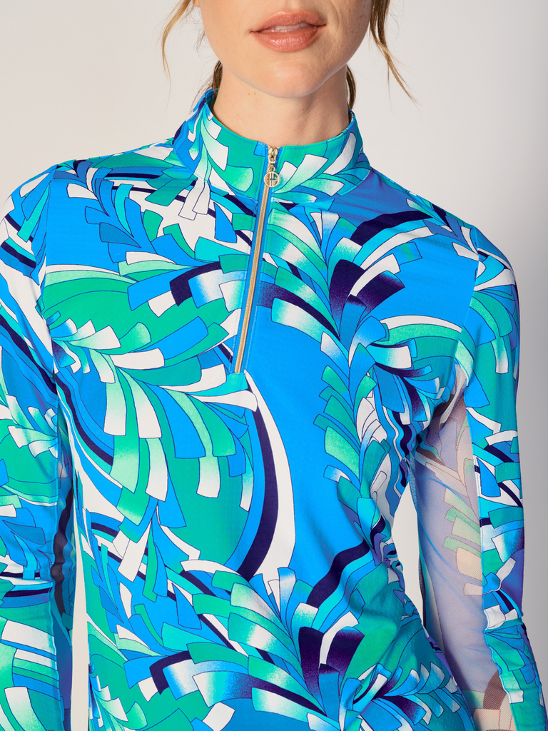 A close-up of a woman adjusting the collar of her G Lifestyle Quarter Zip Sun Protection Top in St Barths Blue - abstract print of the Spring Summer 2024 Collection. The top is characterized by its high neckline and a prominent quarter-zip with a circular zipper pull bearing the brand’s logo. The zipper is gold-toned, adding a touch of luxury to the top. The fabric has a texture, suggesting stretch and comfort, designed to offer sun protection. 