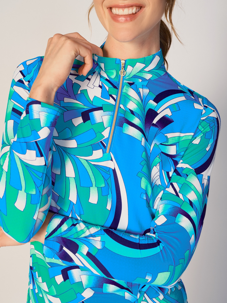 A close-up of a woman adjusting the collar of her G Lifestyle Quarter Zip Sun Protection Top in St Barths Blue - abstract print of the Spring Summer 2024 Collection. The top is characterized by its high neckline and a prominent quarter-zip with a circular zipper pull bearing the brand’s logo. The zipper is gold-toned, adding a touch of luxury to the top. The fabric has a texture, suggesting stretch and comfort, designed to offer sun protection. 