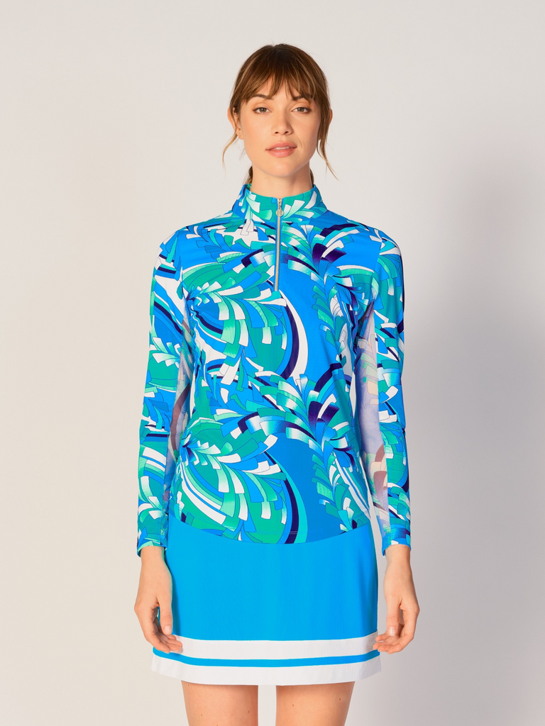 A woman models the G Lifestyle Quarter Zip Sun Protection Top in St Barths Blue - abstract print of the Spring Summer 2024 Collection. The top has long sleeves, an adjustable mock neck collar with a gold quarter zip, logo placed on the right wrist. The fabric appears to have a lightweight, subtle texture, that likely offers menthol cooling, moisture wicking, breathability, sun safety and comfort. Suitable for golf, tennis, padel tennis, pickleball or even cycling.