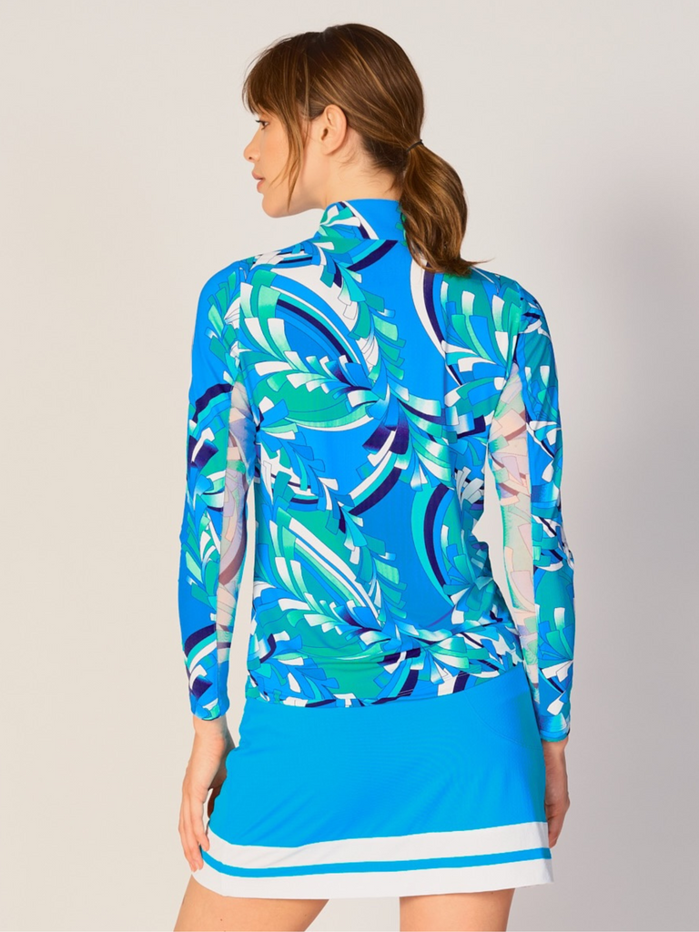 A woman is showcasing the back of the G Lifestyle Quarter Zip Sun Protection Top in St Barths Blue -abstract print of the Spring Summer 2024 Collection. The top is tailored with a form-fitting silhouette, complete with long sleeves and a high collar for maximum sun protection. Notably, it includes smooth mesh underarm inserts, which are seamlessly integrated for enhanced breathability, ensuring comfort during active use. 