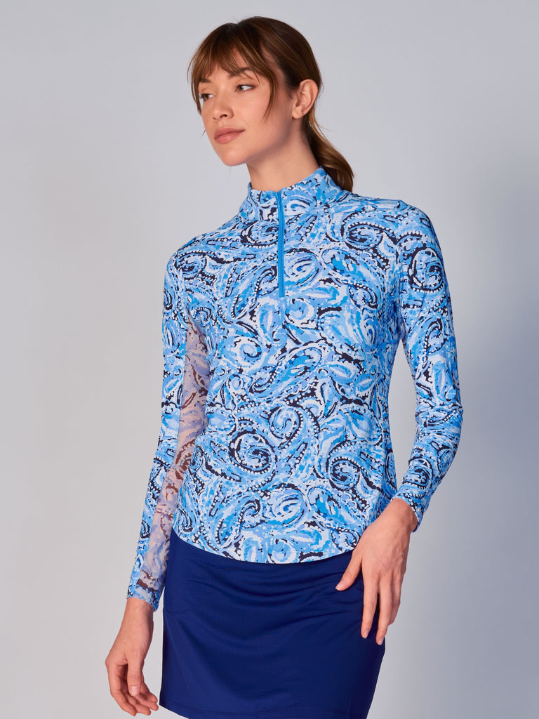 A woman wears the G Lifestyle Quarter Zip Sun Protection Top in Mandala Blue – paisley print of the Spring Summer 2024 Collection. The top has long sleeves, an adjustable mock neck collar with a white quarter zip. The fabric appears to have a lightweight, subtle texture, that likely offers menthol cooling, moisture wicking, breathability, sun safety and comfort. Suitable for golf, tennis, padel tennis, pickleball or even cycling.