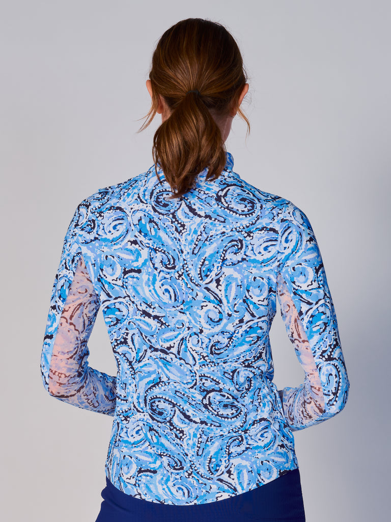A woman is showcasing the back of the G Lifestyle Quarter Zip Sun Protection Top in Mandala Blue – paisley print of the Spring Summer 2024 Collection. The top is tailored with a form-fitting silhouette, complete with long sleeves and a high collar for maximum sun protection. Notably, it includes smooth mesh underarm inserts, which are seamlessly integrated for enhanced breathability, ensuring comfort during active use. 