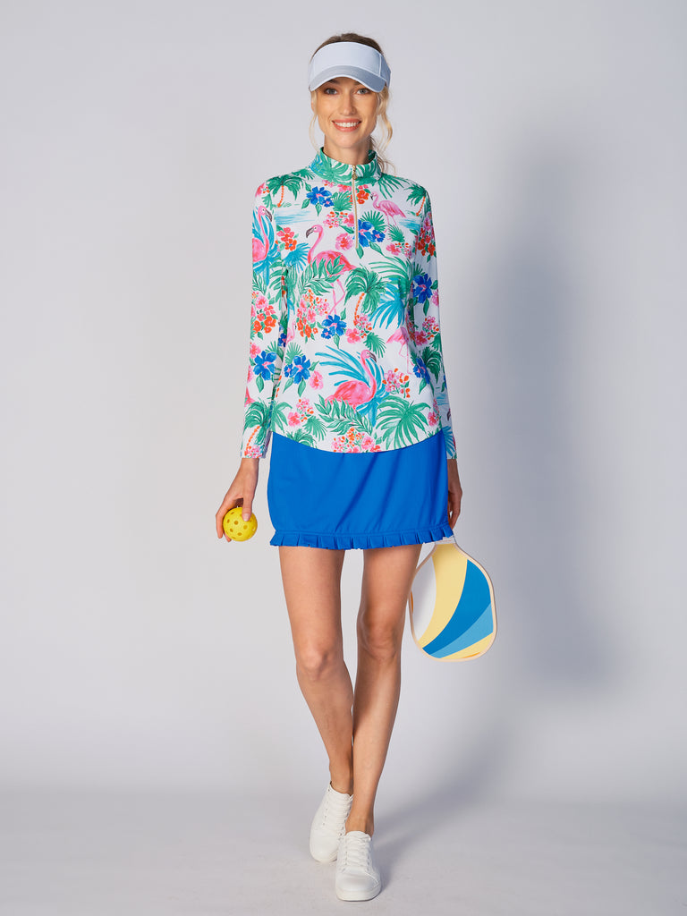 A woman stands, holding a pickleball padel in her hand, showcasing a G Lifestyle Quarter Zip Sun Protection Top in Flamingo - tropical print of the Spring Summer 2024 Collection. The top has long sleeves, an adjustable mock neck collar with a gold quarter zip, logo placed on the right wrist. The fabric appears to have a lightweight, subtle texture, that likely offers menthol cooling, moisture wicking, breathability, sun safety and comfort. Suitable for golf, tennis, padel tennis, pickleball or even cycling.