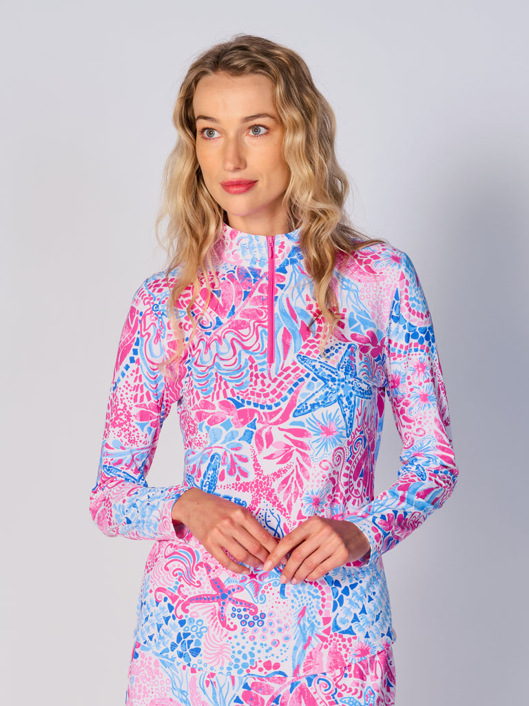 A woman models the G Lifestyle Quarter Zip Sun Protection Top in Starfish Pink – sea-theme print of the Spring Summer 2024 Collection. The top has long sleeves, an adjustable mock neck collar with a flat quarter zip. The fabric appears to have a lightweight, subtle texture, that likely offers menthol cooling, moisture wicking, breathability, sun safety and comfort. Suitable for golf, tennis, padel tennis, pickleball or even cycling.