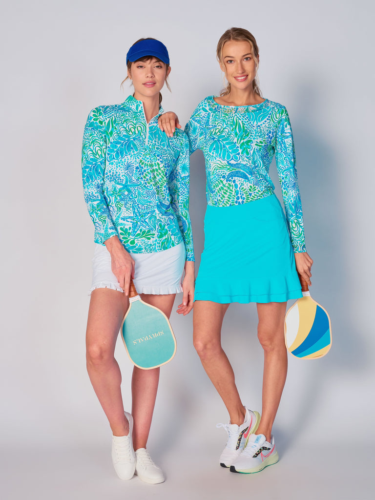 Two women stand side by side, dressed in G Lifestyle Starfish Bright Peri coordinated outfits ready for a game of pickleball. The woman on the left wears G Lifestyle Quarter Zip Sun Protection Top in Starfish Bright Peri and White Ruffle Tennis Skirt. The woman on the right wears G Lifestyle Mesh Block Long Sleeve Top in Starfish Bright Peri and Tiered Ruffle Tennis Skirt in Caribbean Turquoise. Both hold pickleball paddles.