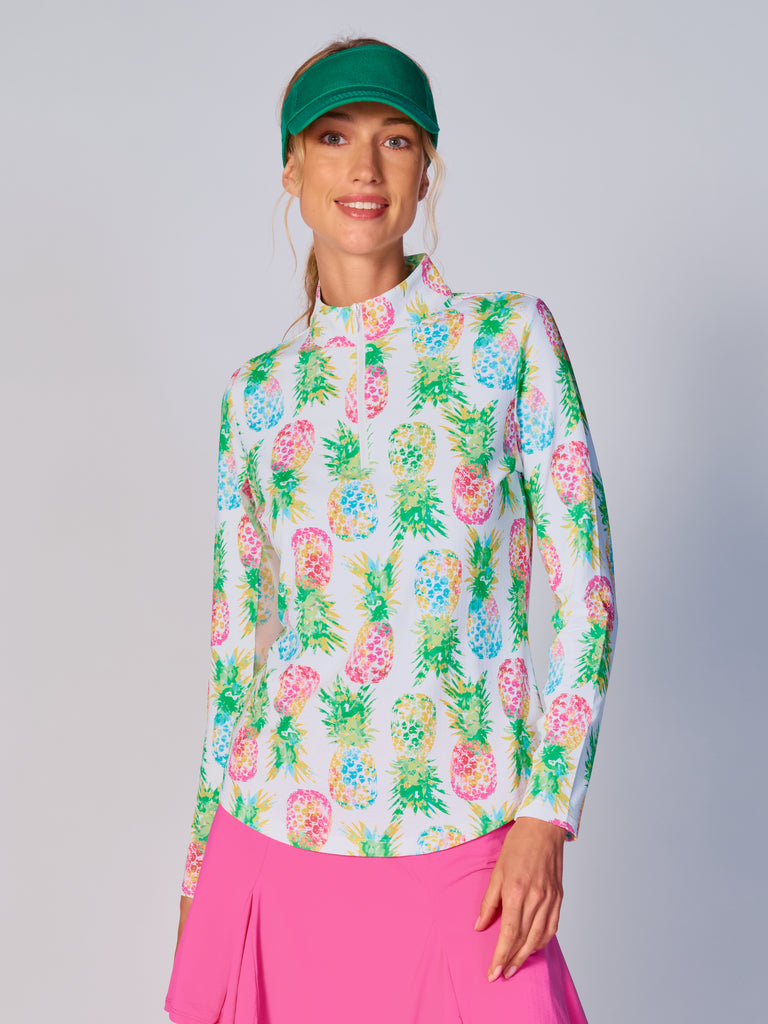 A woman wears the G Lifestyle Quarter Zip Sun Protection Top in Ananas Green - tropical print of the Spring Summer 2024 Collection. The top has long sleeves, an adjustable mock neck collar with a white quarter zip. The fabric appears to have a lightweight, subtle texture, that likely offers menthol cooling, moisture wicking, breathability, sun safety and comfort. Suitable for golf, tennis, padel tennis, pickleball or even cycling.