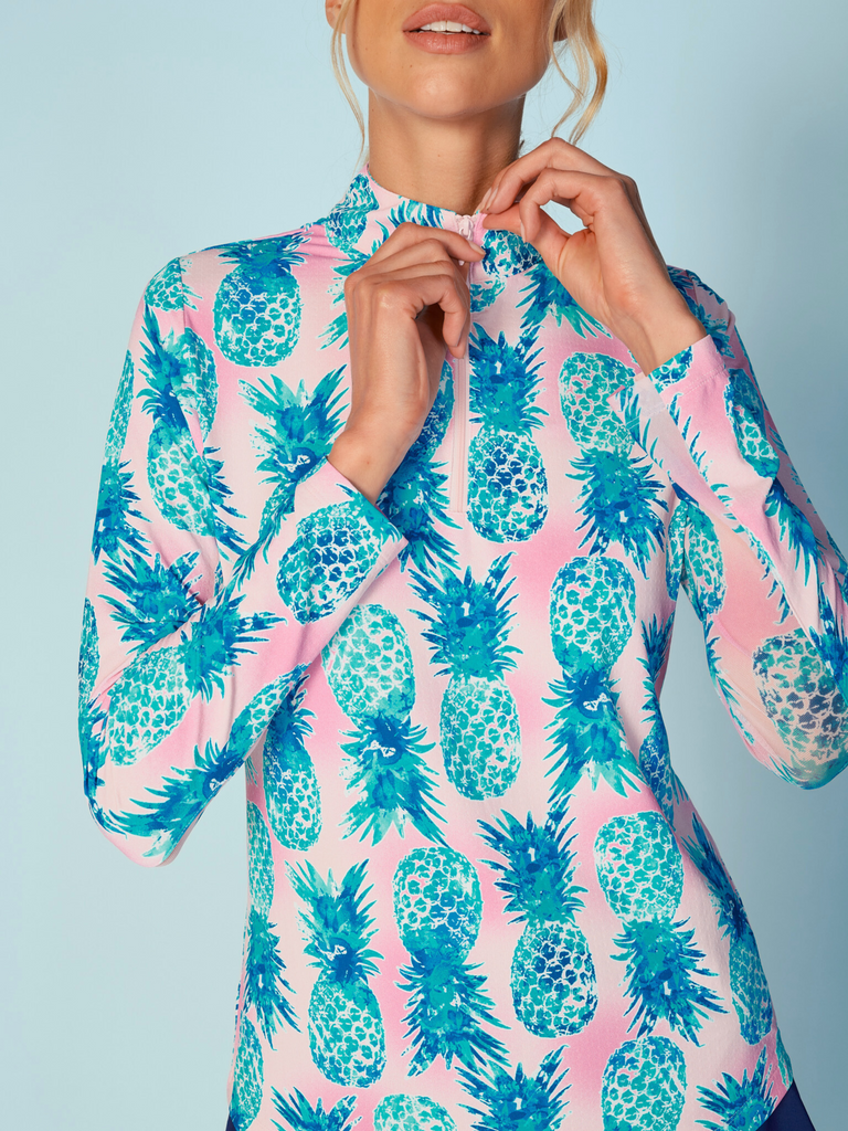 A close-up of a woman adjusting the collar of her G Lifestyle Quarter Zip Sun Protection Top in Ananas Aqua - tropical print of the Spring Summer 2024 Collection. The top is characterized by its high neckline and a flat quarter-zip. The fabric has a texture, suggesting stretch and comfort, designed to offer sun protection. 