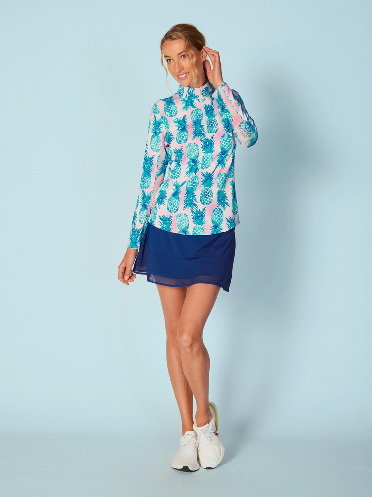 A woman wears the G Lifestyle Quarter Zip Sun Protection Top in Ananas Aqua - tropical print of the Spring Summer 2024 Collection. The top has long sleeves, an adjustable mock neck collar with a white quarter zip. The fabric appears to have a lightweight, subtle texture, that likely offers menthol cooling, moisture wicking, breathability, sun safety and comfort. Suitable for golf, tennis, padel tennis, pickleball or even cycling.
