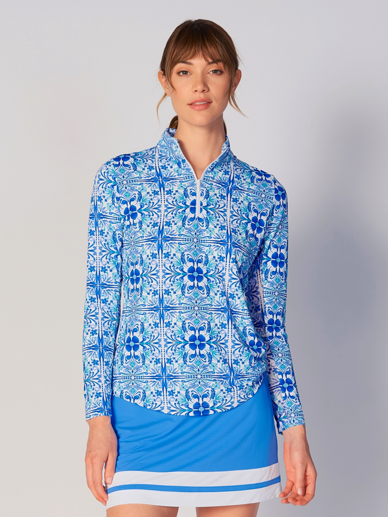 A woman models the G Lifestyle Quarter Zip Sun Protection Top in Blue Tile – symmetrical ornamental print of the Spring Summer 2024 Collection. The top has long sleeves, an adjustable mock neck collar with a flat quarter zip. The fabric appears to have a lightweight, subtle texture, that likely offers menthol cooling, moisture wicking, breathability, sun safety and comfort. Suitable for golf, tennis, padel tennis, pickleball or even cycling.