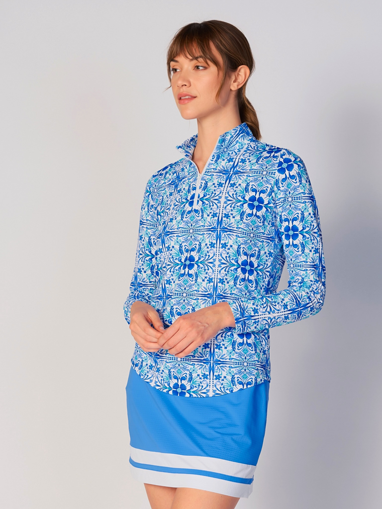 A woman models the G Lifestyle Quarter Zip Sun Protection Top in Blue Tile – symmetrical ornamental print of the Spring Summer 2024 Collection. The top has long sleeves, an adjustable mock neck collar with a flat quarter zip. The fabric appears to have a lightweight, subtle texture, that likely offers menthol cooling, moisture wicking, breathability, sun safety and comfort. Suitable for golf, tennis, padel tennis, pickleball or even cycling.