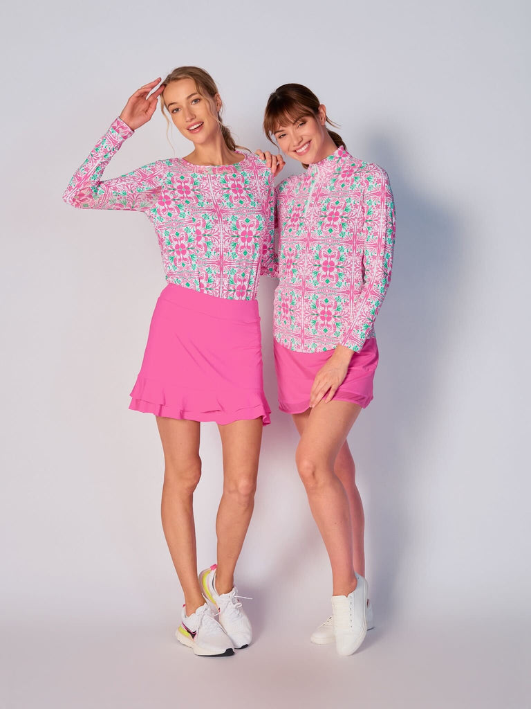 Two women stand side by side, dressed in sporty G Lifestyle Pink Tile coordinated outfits. The woman on the right wears G Lifestyle Quarter Zip Sun Protection Top in Pink Tile and Hot Pink Mesh Block Tennis Skirt. The woman on the right wears G Lifestyle Mesh Block Long Sleeve Top in Pink Tile and Tiered Ruffle Tennis Skirt in Hot Pink. Both women are smiling.