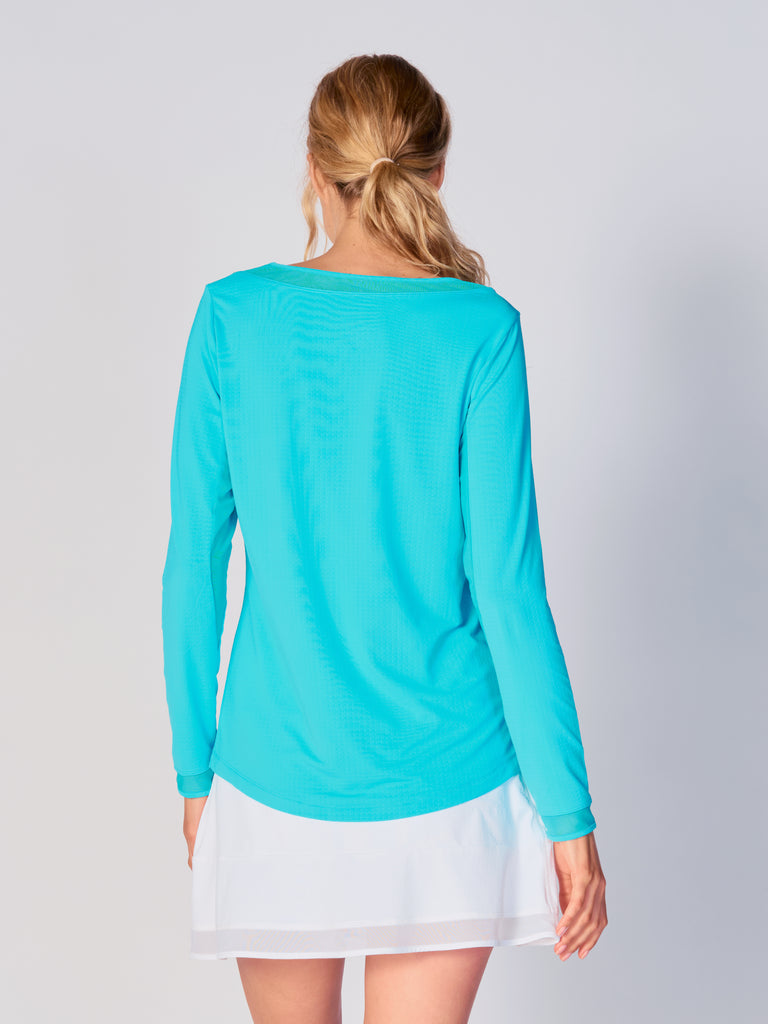A woman is showcasing the back of the G Lifestyle Mesh Block Long Sleeve Top in Caribbean Turquoise. The top is tailored with a snug fit silhouette, complete with long sleeves and round neckline with mesh details. Notably, it includes smooth mesh underarm inserts, which are seamlessly integrated for enhanced breathability, ensuring comfort during active use. 
