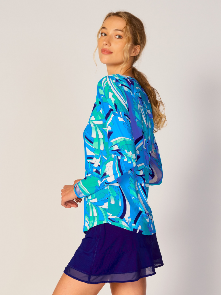 A woman is showcasing the side of the G Lifestyle Mesh Block Long Sleeve Top in St Barths Blue - abstract print of the Spring Summer 2024 Collection. The top is designed with long sleeves, mesh underarm inserts for breathability, and features a round neckline with texture mesh block detailing around it and sleeve hems. The fabric appears to be lightweight, movement-friendly, with subtle texture. Suitable for various outdoor athletic activities such as golf, tennis, padel tennis, pickleball or even cycling.