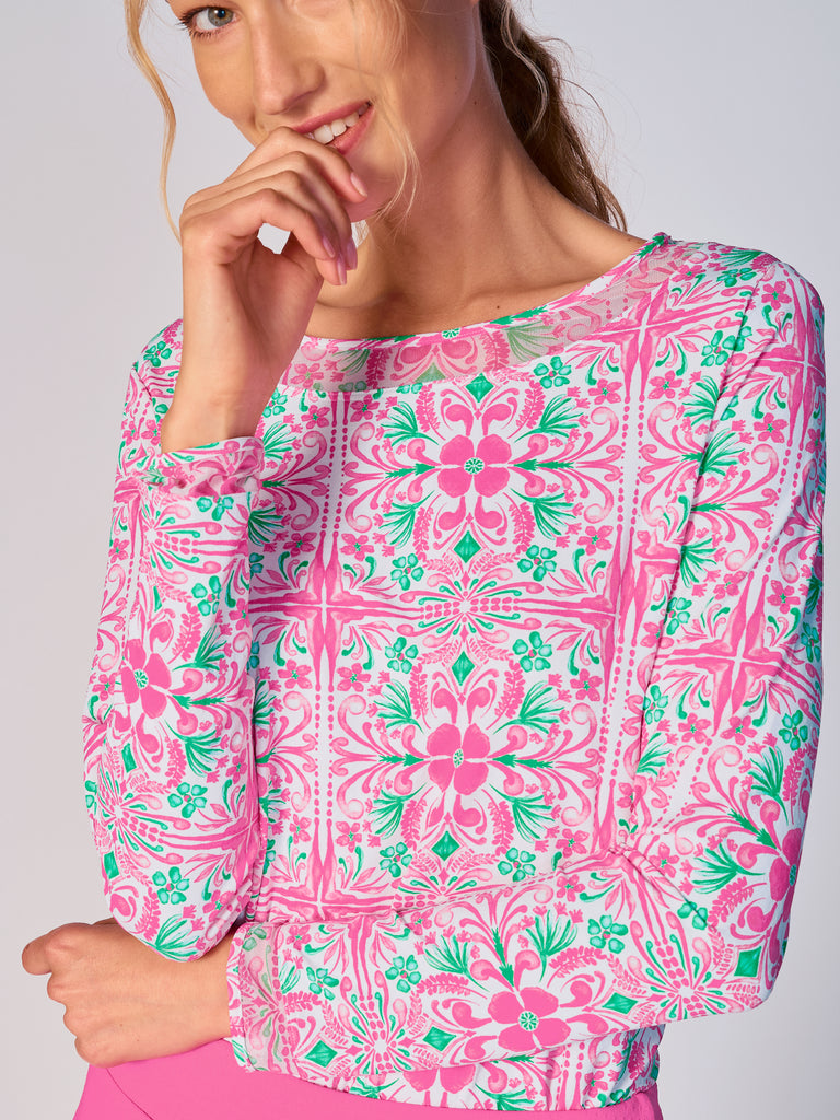 A woman wears G Lifestyle Mesh Block Long Sleeve Top in Pink Tile – symmetrical ornamental print of the Spring Summer 2024 Collection. The top is designed with long sleeves, mesh underarm inserts for breathability, and features a round neckline with texture mesh block detailing around it and sleeve hems. The fabric appears to be lightweight, movement-friendly, with subtle texture. Suitable for various outdoor athletic activities such as golf, tennis, padel tennis, pickleball or even cycling.