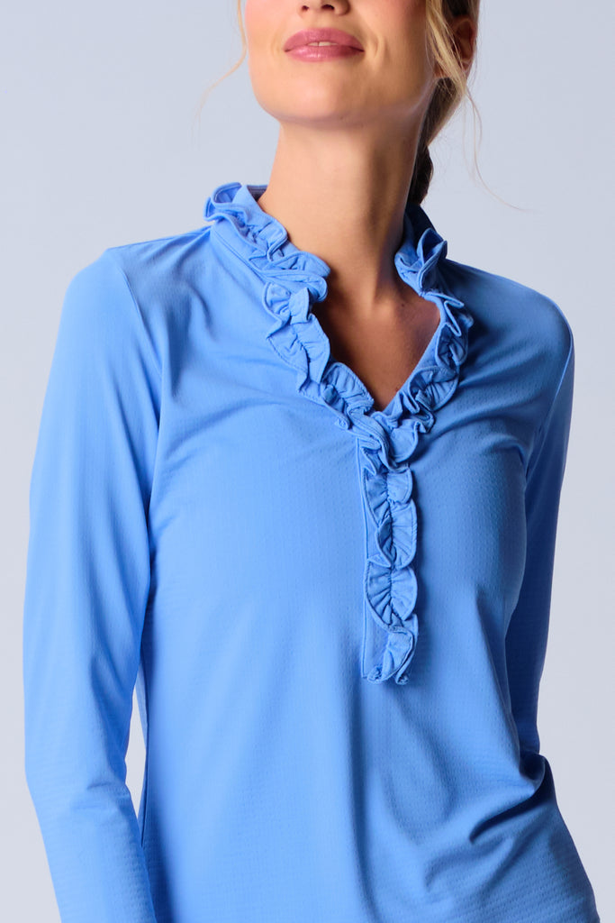 A woman wears G Lifestyle UPF 50+ Ruffle V Neck Top in Bright Peri. The top is designed with long sleeves, mesh underarm inserts for breathability, and features a delicately ruffled V-neckline adding feminine touch to the sporty style. The fabric appears to be lightweight, movement-friendly, with subtle texture. Suitable for various outdoor athletic activities such as golf, tennis, padel tennis, pickleball or even cycling.
