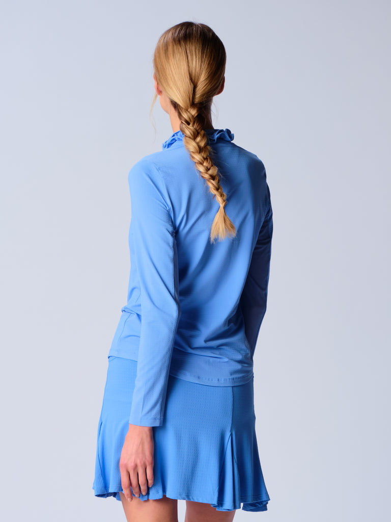 A woman is showcasing the back of the G Lifestyle UPF 50+ Ruffle V Neck Top in Bright Peri. The top is tailored with a snug fit silhouette, complete with long sleeves and a v-neckline with ruffle details. Notably, it includes smooth mesh underarm inserts, which are seamlessly integrated for enhanced breathability, ensuring comfort during active use. 