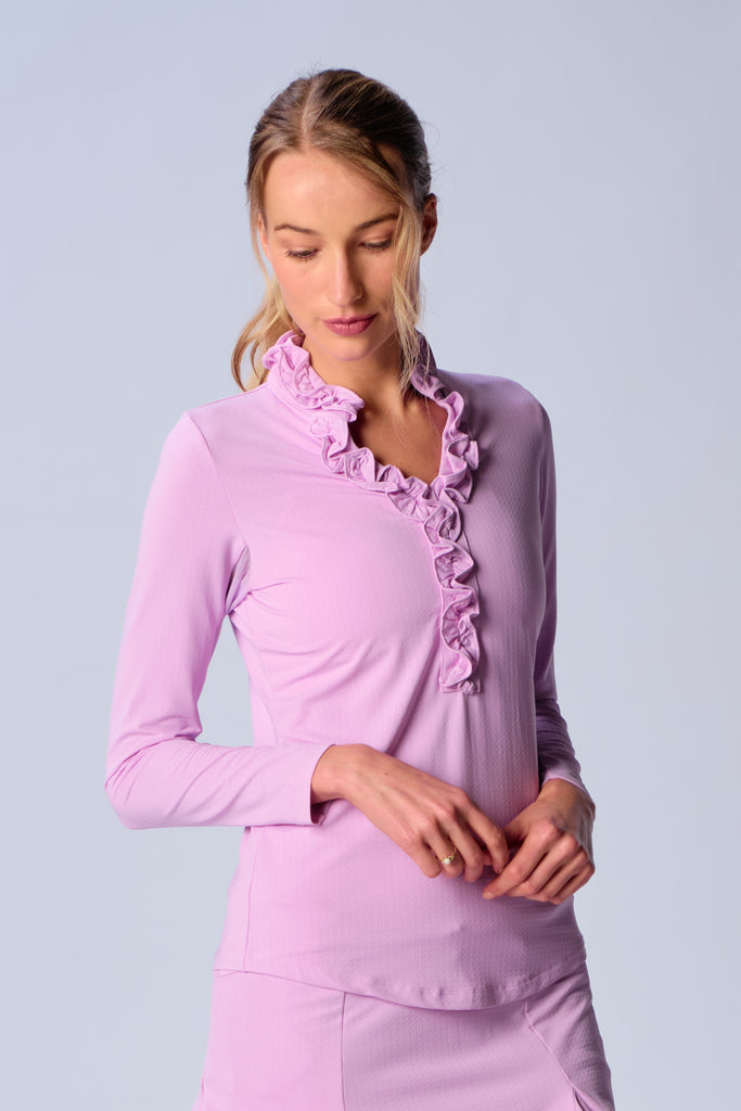 A woman wears G Lifestyle UPF 50+ Ruffle V Neck Top in Lavender. The top is designed with long sleeves, mesh underarm inserts for breathability, and features a delicately ruffled V-neckline adding feminine touch to the sporty style. The fabric appears to be lightweight, movement-friendly, with subtle texture. Suitable for various outdoor athletic activities such as golf, tennis, padel tennis, pickleball or even cycling.