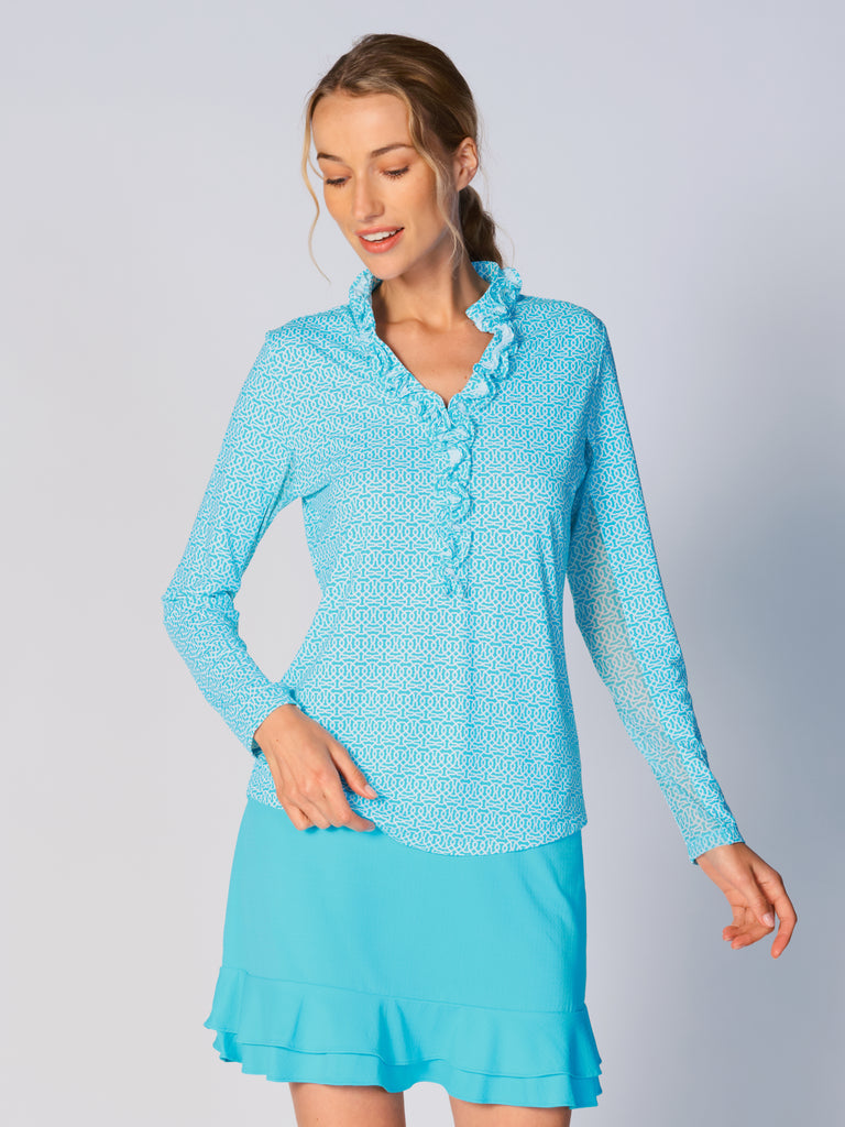A woman wears G Lifestyle UPF 50+ Ruffle V Neck Top in Cubic Caribbean Turquoise - geometric print of the Spring Summer 2024 Collection. The top is designed with long sleeves, mesh underarm inserts for breathability, and features a delicately ruffled V-neckline adding feminine touch to the sporty style. The fabric appears to be lightweight, movement-friendly, with subtle texture. Suitable for various outdoor athletic activities such as golf, tennis, padel tennis, pickleball or even cycling.
