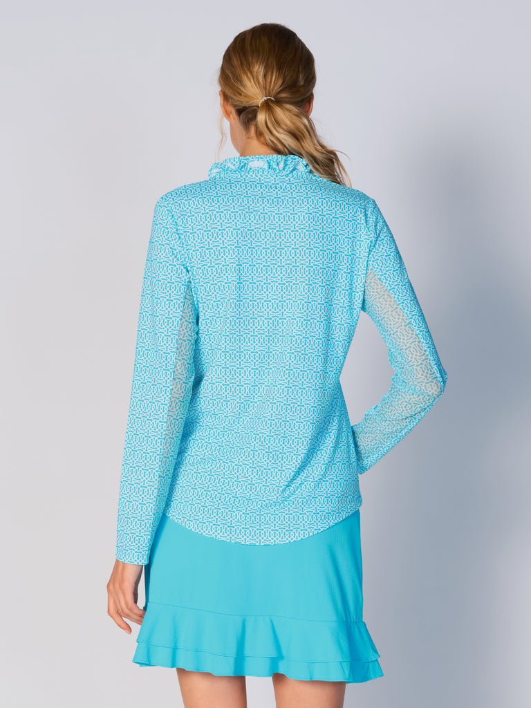 A woman is showcasing the back of the G Lifestyle UPF 50+ Ruffle V Neck Top in Cubic Caribbean Turquoise - geometric print of the Spring Summer 2024 Collection. The top is tailored with a snug fit silhouette, complete with long sleeves and a v-neckline with ruffle details. Notably, it includes smooth mesh underarm inserts, which are seamlessly integrated for enhanced breathability, ensuring comfort during active use. 