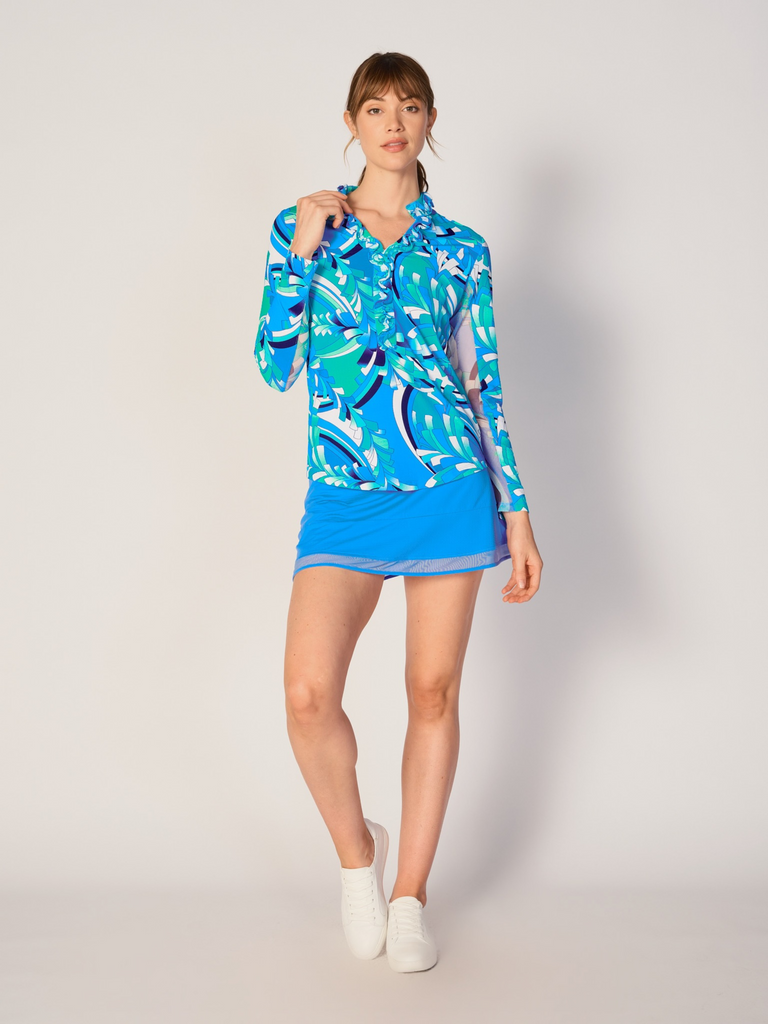A woman wears G Lifestyle UPF 50+ Ruffle V Neck Top in St Barths Blue - abstract print of the Spring Summer 2024 Collection. The top is designed with long sleeves, mesh underarm inserts for breathability, and features a delicately ruffled V-neckline adding feminine touch to the sporty style. The fabric appears to be lightweight, movement-friendly, with subtle texture. Suitable for various outdoor athletic activities such as golf, tennis, padel tennis, pickleball or even cycling.