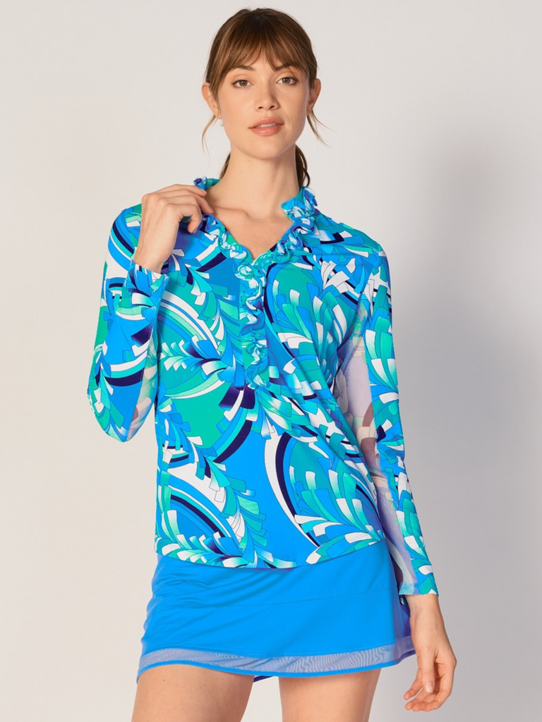 A woman wears G Lifestyle UPF 50+ Ruffle V Neck Top in St Barths Blue - abstract print of the Spring Summer 2024 Collection. The top is designed with long sleeves, mesh underarm inserts for breathability, and features a delicately ruffled V-neckline adding feminine touch to the sporty style. The fabric appears to be lightweight, movement-friendly, with subtle texture. Suitable for various outdoor athletic activities such as golf, tennis, padel tennis, pickleball or even cycling.