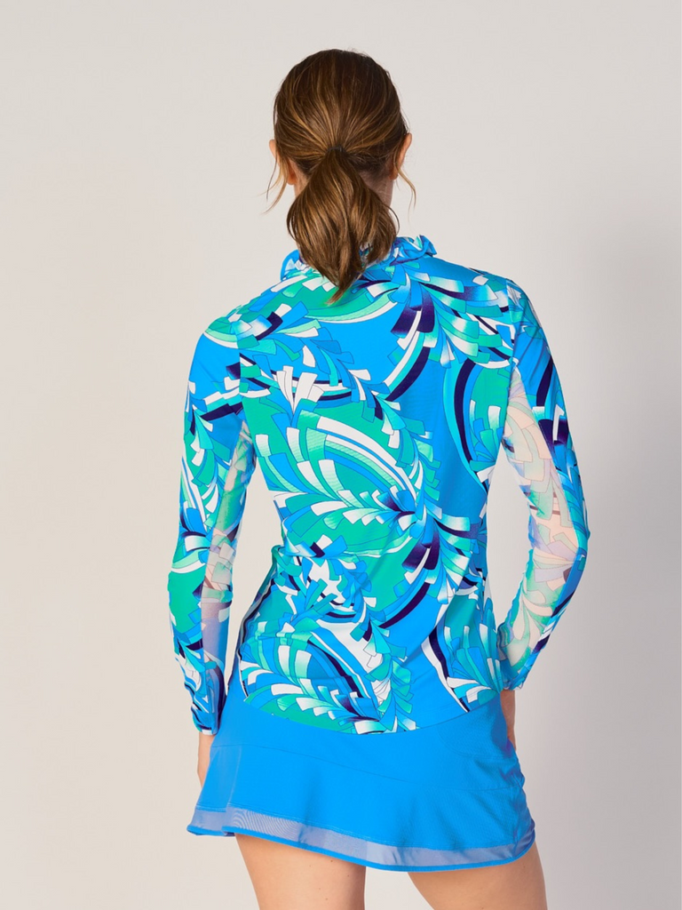A woman is showcasing the back of the G Lifestyle UPF 50+ Ruffle V Neck Top in St Barths Blue - abstract print of the Spring Summer 2024 Collection. The top is tailored with a snug fit silhouette, complete with long sleeves and a v-neckline with ruffle details. Notably, it includes smooth mesh underarm inserts, which are seamlessly integrated for enhanced breathability, ensuring comfort during active use. 