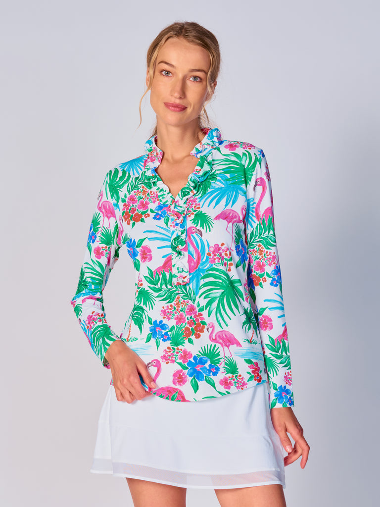 A woman wears G Lifestyle UPF 50+ Ruffle V Neck Top in Flamingo - tropical print of the Spring Summer 2024 Collection. The top is designed with long sleeves, mesh underarm inserts for breathability, and features a delicately ruffled V-neckline adding feminine touch to the sporty style. The fabric appears to be lightweight, movement-friendly, with subtle texture. Suitable for various outdoor athletic activities such as golf, tennis, padel tennis, pickleball or even cycling.