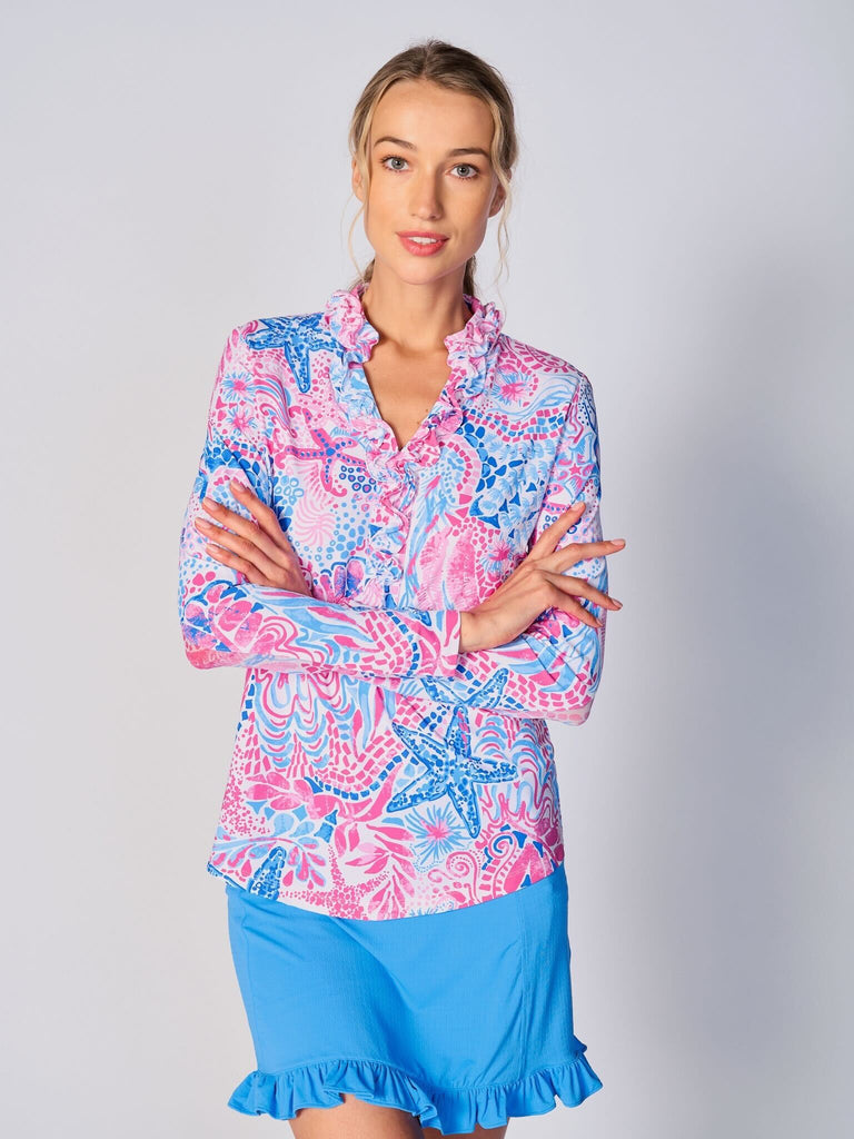 A woman wears G Lifestyle UPF 50+ Ruffle V Neck Top in Starfish Pink – sea-theme print of the Spring Summer 2024 Collection. The top is designed with long sleeves, mesh underarm inserts for breathability, and features a delicately ruffled V-neckline adding feminine touch to the sporty style. The fabric appears to be lightweight, movement-friendly, with subtle texture. Suitable for various outdoor athletic activities such as golf, tennis, padel tennis, pickleball or even cycling.