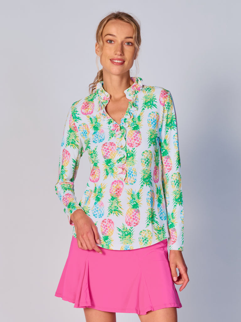 A woman wears G Lifestyle UPF 50+ Ruffle V Neck Top in Ananas Green - tropical print of the Spring Summer 2024 Collection. The top is designed with long sleeves, mesh underarm inserts for breathability, and features a delicately ruffled V-neckline adding feminine touch to the sporty style. The fabric appears to be lightweight, movement-friendly, with subtle texture. Suitable for various outdoor athletic activities such as golf, tennis, padel tennis, pickleball or even cycling.