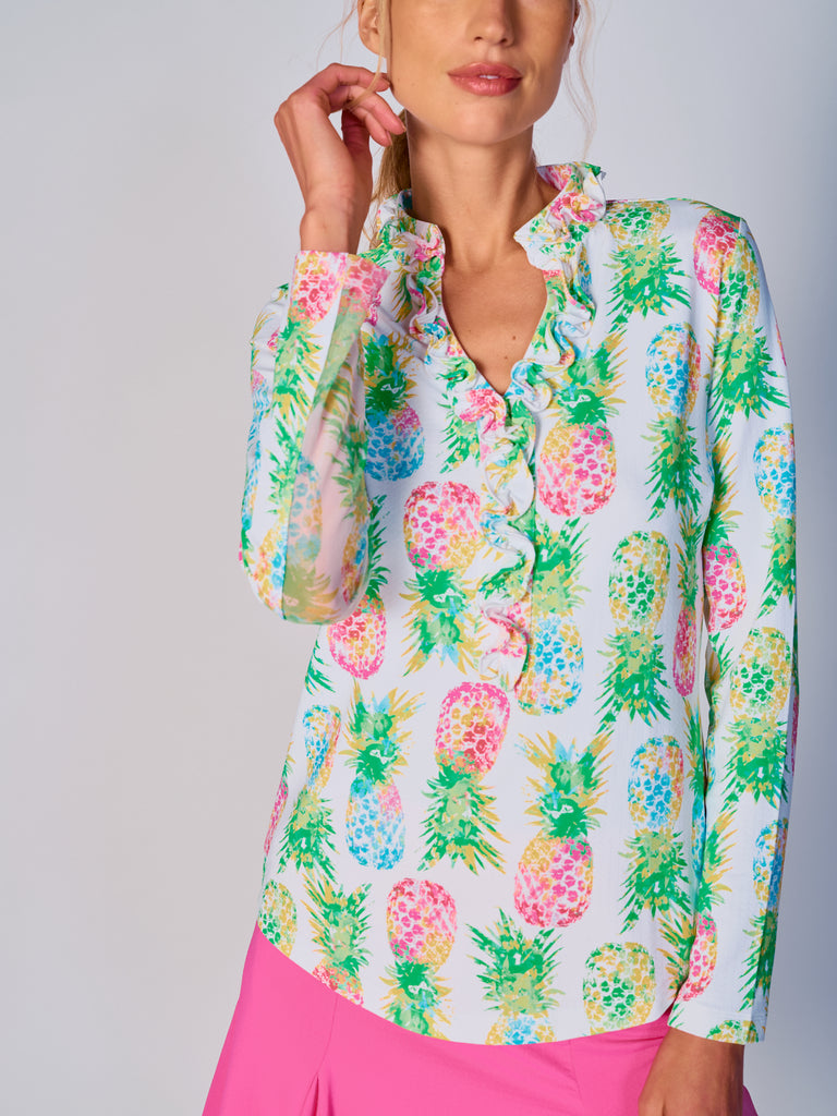 A woman wears G Lifestyle UPF 50+ Ruffle V Neck Top in Ananas Green - tropical print of the Spring Summer 2024 Collection. The top is designed with long sleeves, mesh underarm inserts for breathability, and features a delicately ruffled V-neckline adding feminine touch to the sporty style. The fabric appears to be lightweight, movement-friendly, with subtle texture. Suitable for various outdoor athletic activities such as golf, tennis, padel tennis, pickleball or even cycling.
