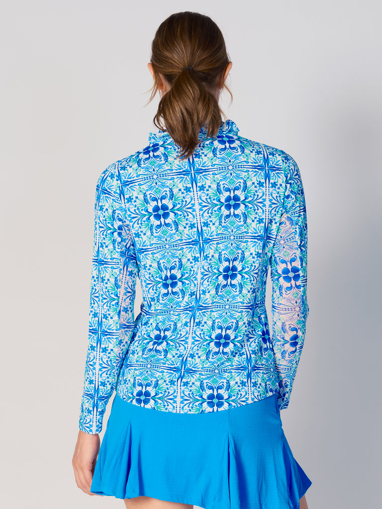 A woman is showcasing the back of the G Lifestyle UPF 50+ Ruffle V Neck Top in Blue Tile – symmetrical ornamental print of the Spring Summer 2024 Collection. The top is tailored with a snug fit silhouette, complete with long sleeves and a v-neckline with ruffle details. Notably, it includes smooth mesh underarm inserts, which are seamlessly integrated for enhanced breathability, ensuring comfort during active use. 